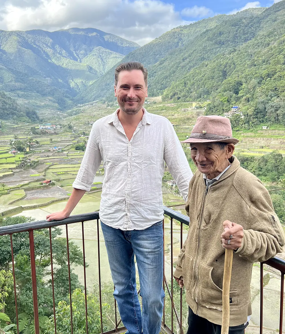 The author of the e-book stands at a viewpoint next to an elderly Filipino man. Behind them, amphitheater-like stone terraces of Batad roll down the mountain valley.