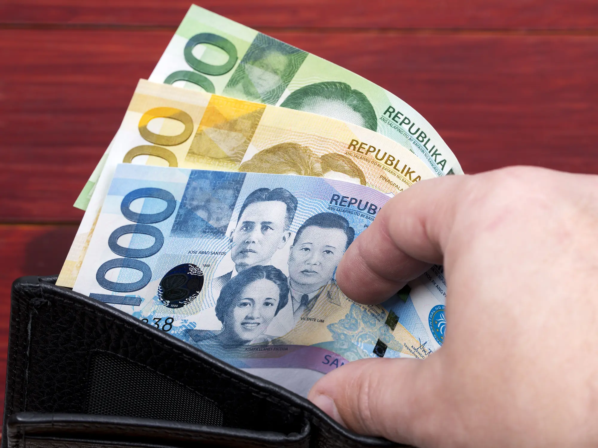 A black leather wallet containing three Philippine Peso bills a blue 1,000-piso note, a yellow 500-piso note, and a green 200-piso note.